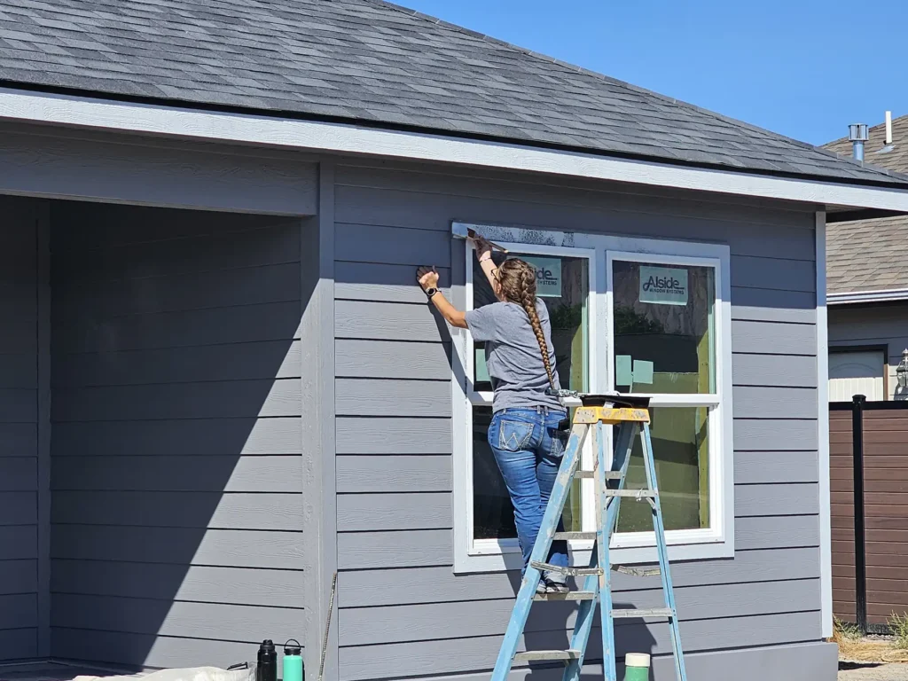 A woman stands on a ladder and paints the outdoor trim of a window on a new home.